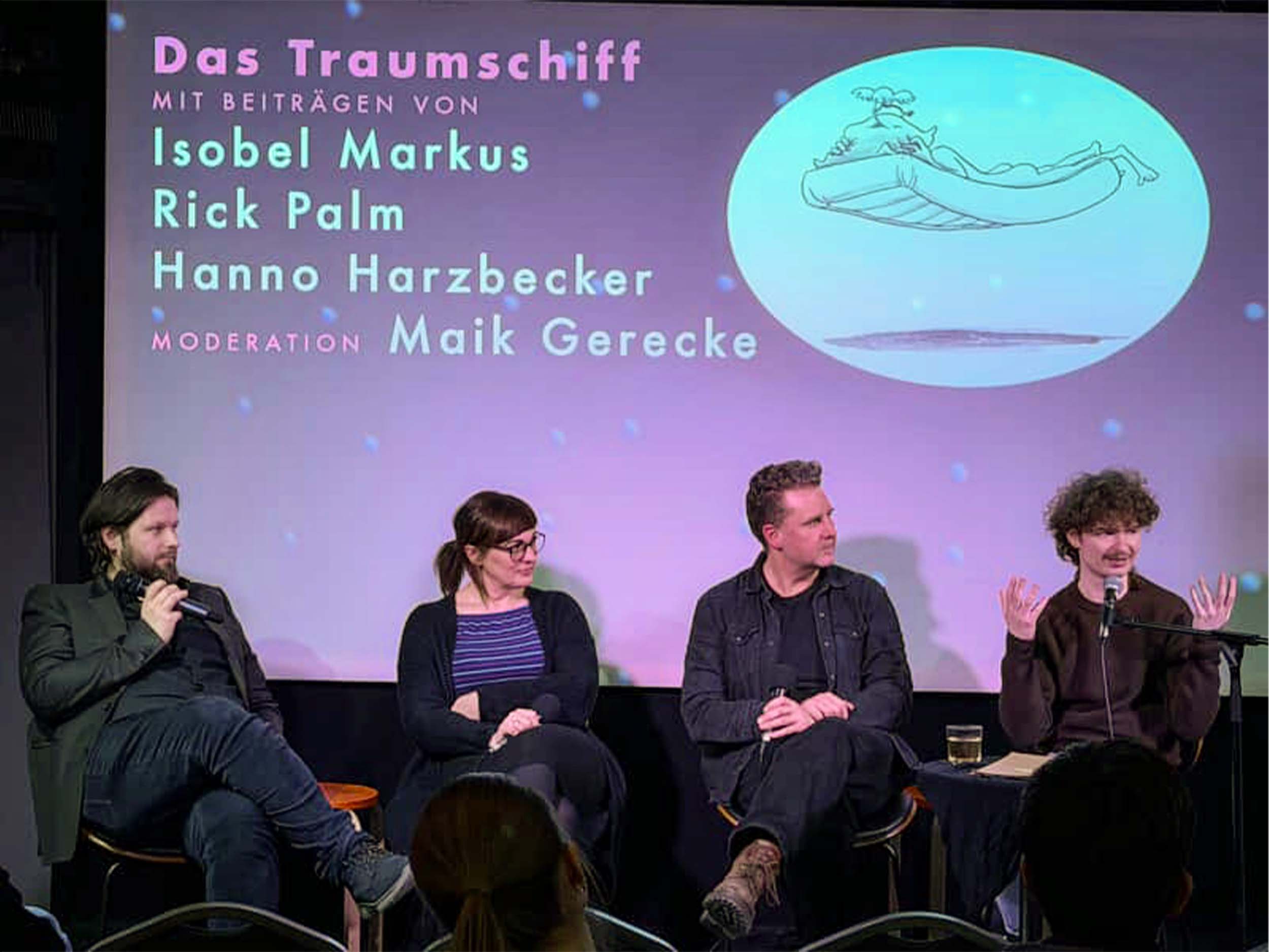 Foto von einer Bühne. Vier Menschen sitzen auf Stühlen und schauen nach rechts. Der Mensch ganz rechts gestikuliert und spricht in ein Mikrofon. Im Hintergrund ist eine Leinwand mit Grafik und Text. Die Menschen sind von links nach rechts: Maik Gerecke, Isobel Markus, Hanno Harzbecker, Rick Palm. Auf der Leinwand steht "Das Traumschiff. Mit Beiträgen von Isobel Markus, Rick Palm, Hanno Harzbecker. Moderation: Maik Gerecke". Daneben ist eine Zeichnung zu sehen, auf der ein Mensch nackt auf einer fliegenden Matratze liegt und noch weiterschlafen möchte.