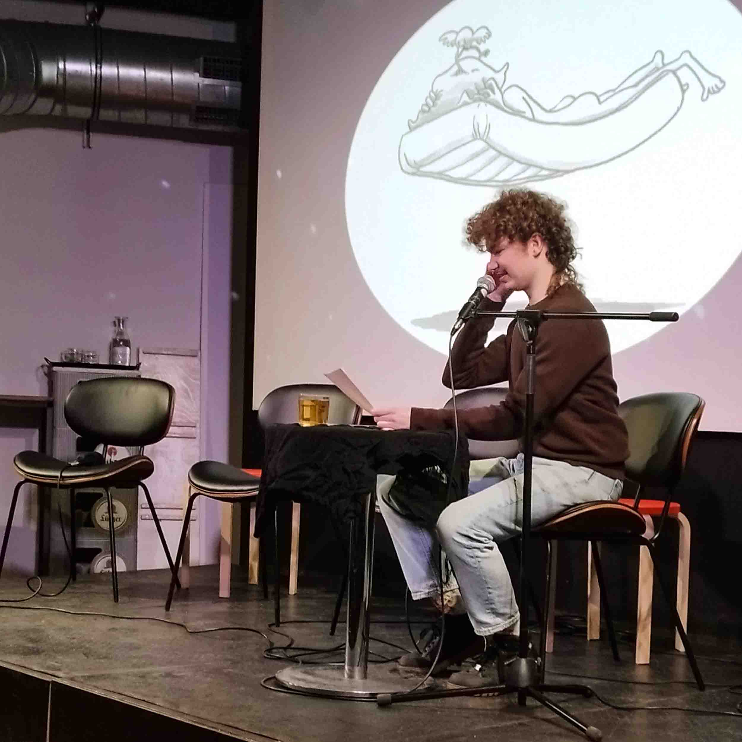 Foto von einer Bühne. Ein Mensch sitzt auf einem Stuhl an einem Tisch und liest vor. Der Mensch ist Rick Palm. Rick trägt einen braunen Pullover und eine helle Jeans. Im Hintergrund ist eine Leinwand mit einer Grafik. Die Grafik ist eine Zeichnung, auf der ein Mensch nackt auf einer fliegenden Matratze liegt und noch weiterschlafen möchte. Er hält sich das Kissen über den Kopf, aber ein Vogel versucht ihn zu wecken.