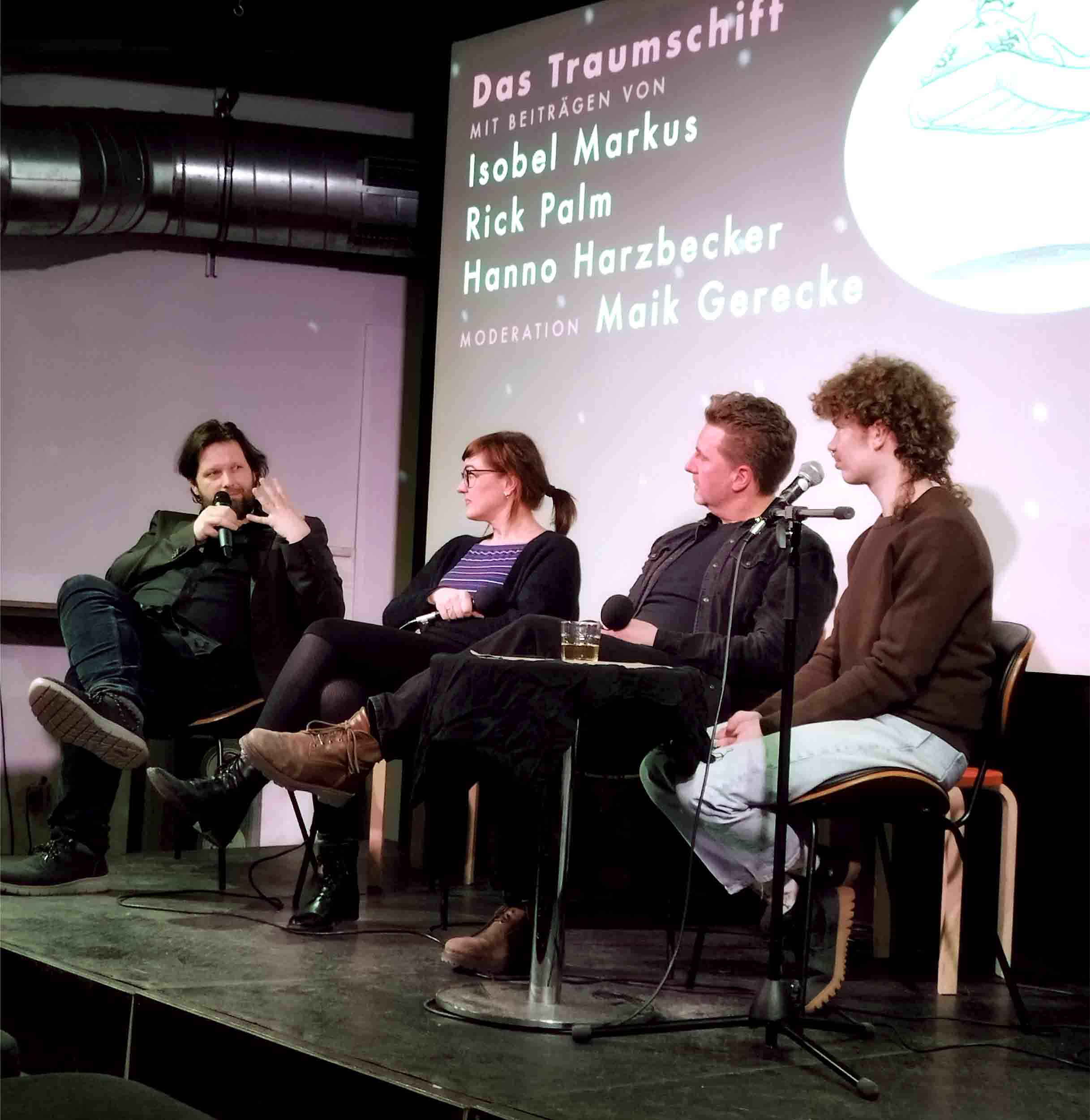 Foto von einer Bühne. Vier Menschen sitzen auf Stühlen. Ein Mensch ganz links gestikuliert und spricht in ein Mikrofon, die anderen drei schauen zu ihm. Im Hintergrund ist eine Leinwand mit Grafik und Text. Die Menschen sind von links nach rechts: Maik Gerecke, Isobel Markus, Hanno Harzbecker, Rick Palm. Auf der Leinwand steht "Das Traumschiff. Mit Beiträgen von Isobel Markus, Rick Palm, Hanno Harzbecker. Moderation: Maik Gerecke". Daneben ist eine Zeichnung zu sehen, auf der ein Mensch nackt auf einer fliegenden Matratze liegt und noch weiterschlafen möchte.